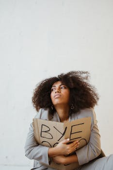 African woman in gray suit holding 'Bye 2020' sign, looking thoughtful.
