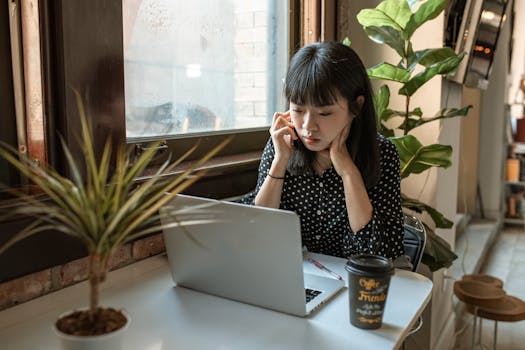 Asian woman focused on work in a modern office setting with laptop and coffee.
