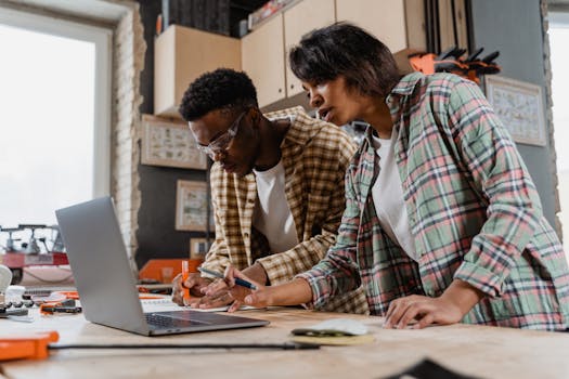 Two people focused on a woodworking project in a workshop, using a laptop and tools.