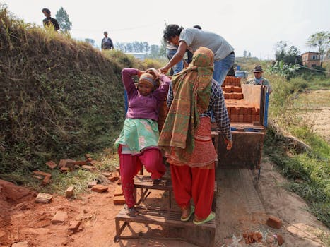 Workers load bricks onto a truck on an unpaved road in the countryside, highlighting manual labor in rural areas.